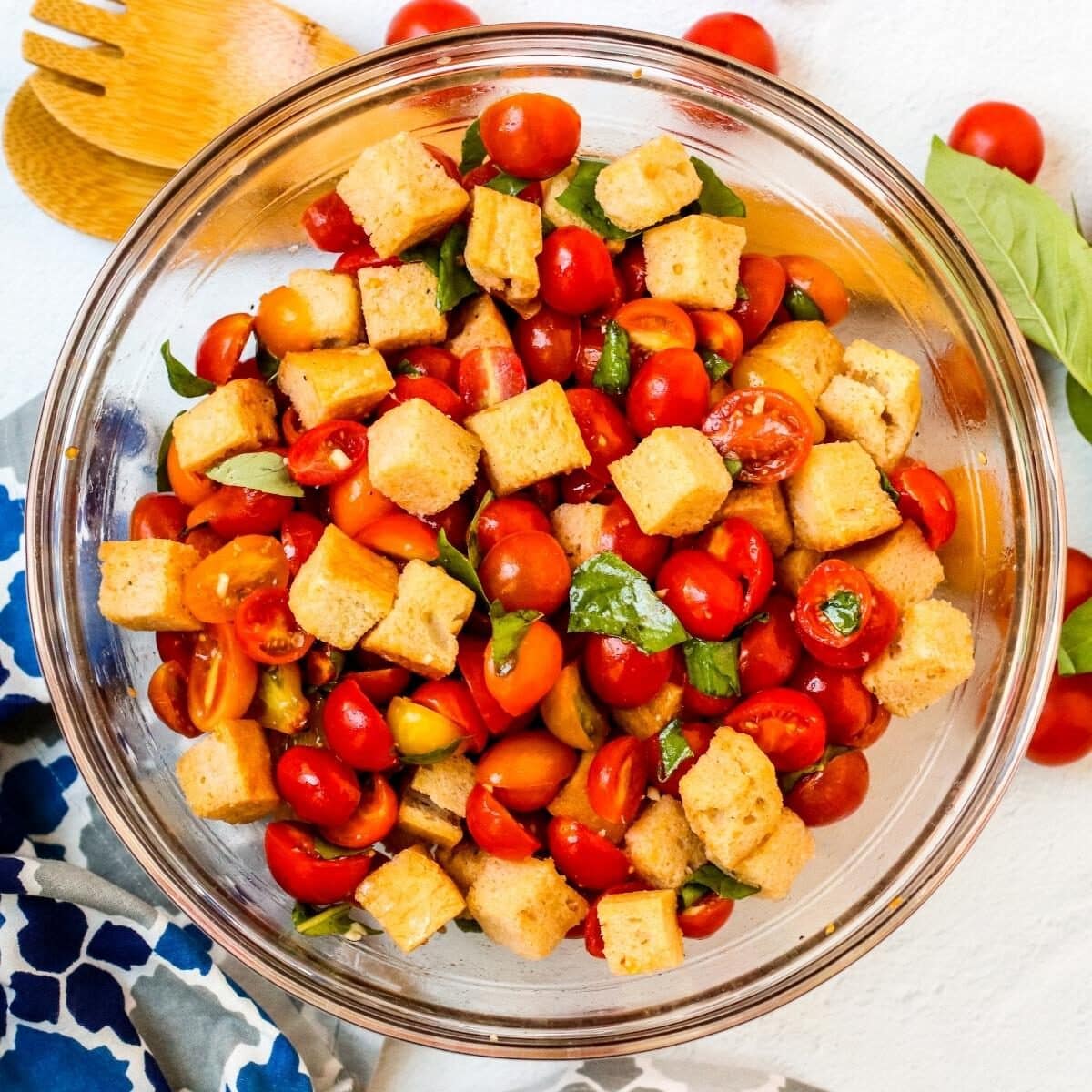 Bowl of tomato and bread salad with serving utensils and a blue pattered napkin on the side.