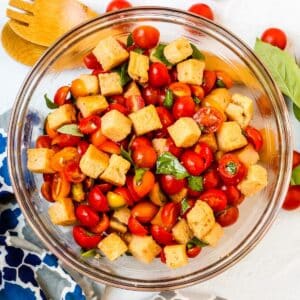 Bowl of tomato and bread salad with serving utensils and a blue pattered napkin on the side.