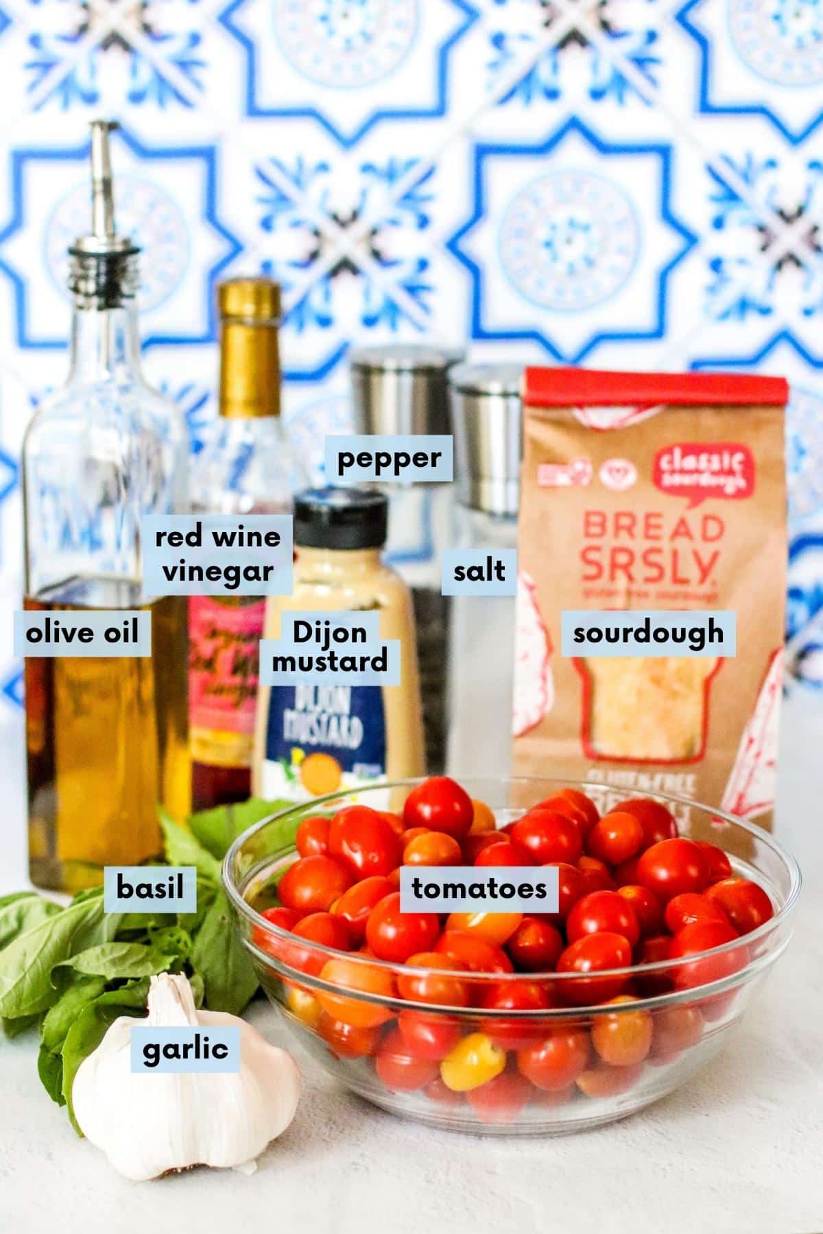 Labeled ingredients needed to make this recipe: olive oil, red wine vinegar, Dijon mustard, pepper, salt, loaf of sourdough bread, large bowl of tomatoes, fresh basil, and a head of garlic.