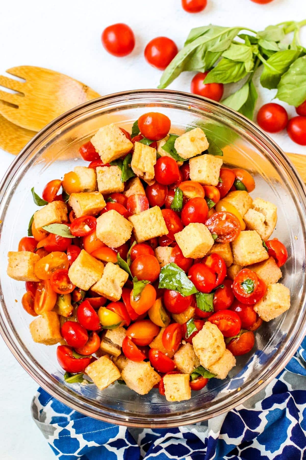 Bread Salad in a glass bowl surrounded by serving utensils, cherry tomatoes, fresh basil, and a blue patterned napkin.