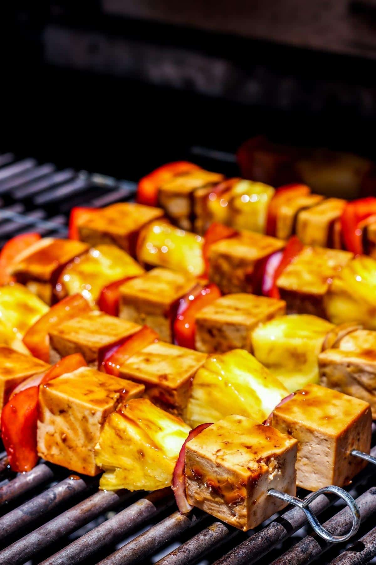 Tofu, pineapple, red bell pepper, and onion on metal skewers cooking on a barbecue grill.