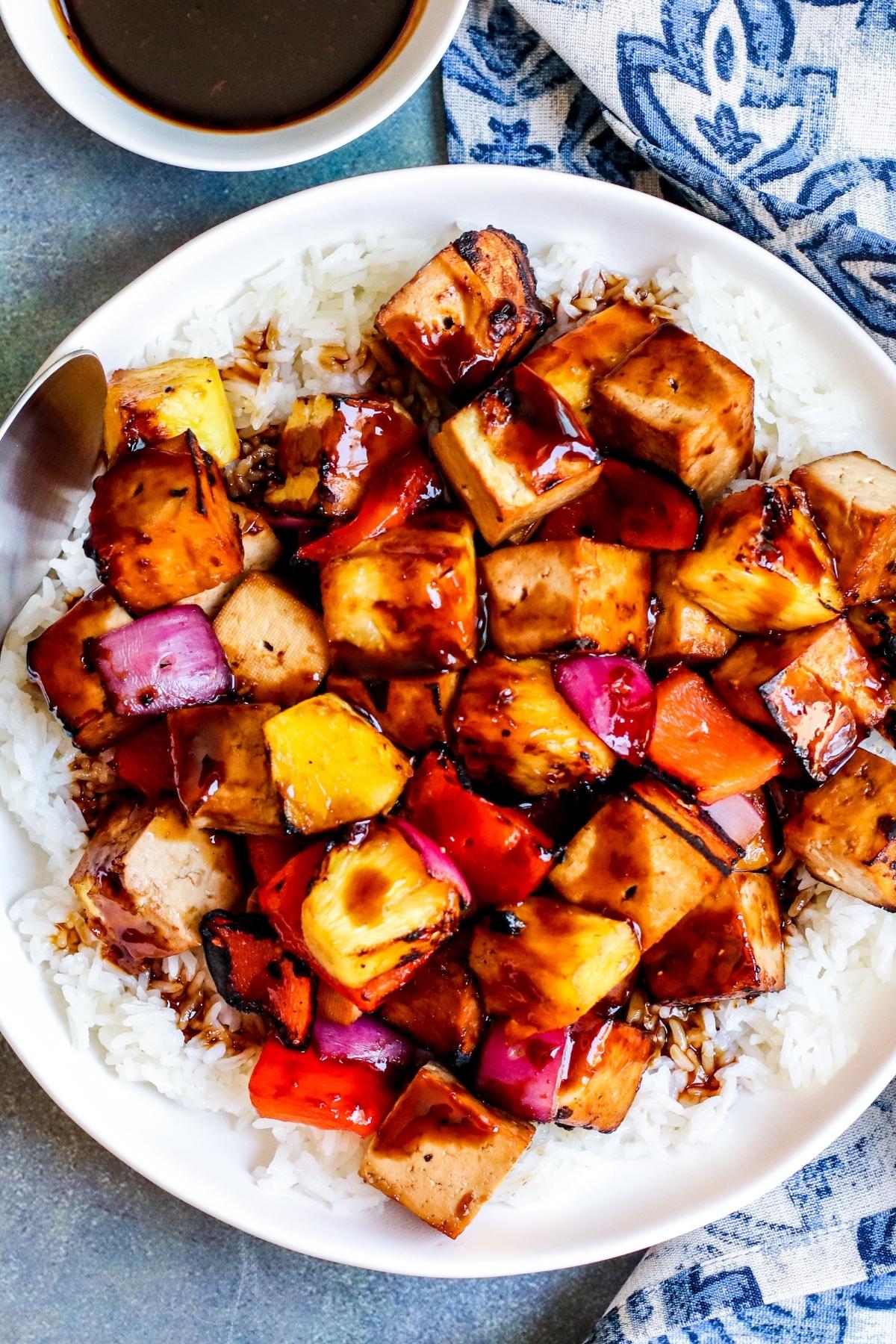 Teriyaki tofu and pineapple on a plate of rice with small bowl of sauce next to it.