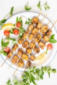 Tofu skewers on a plate with fresh herbs, cherry tomatoes, and lemon.