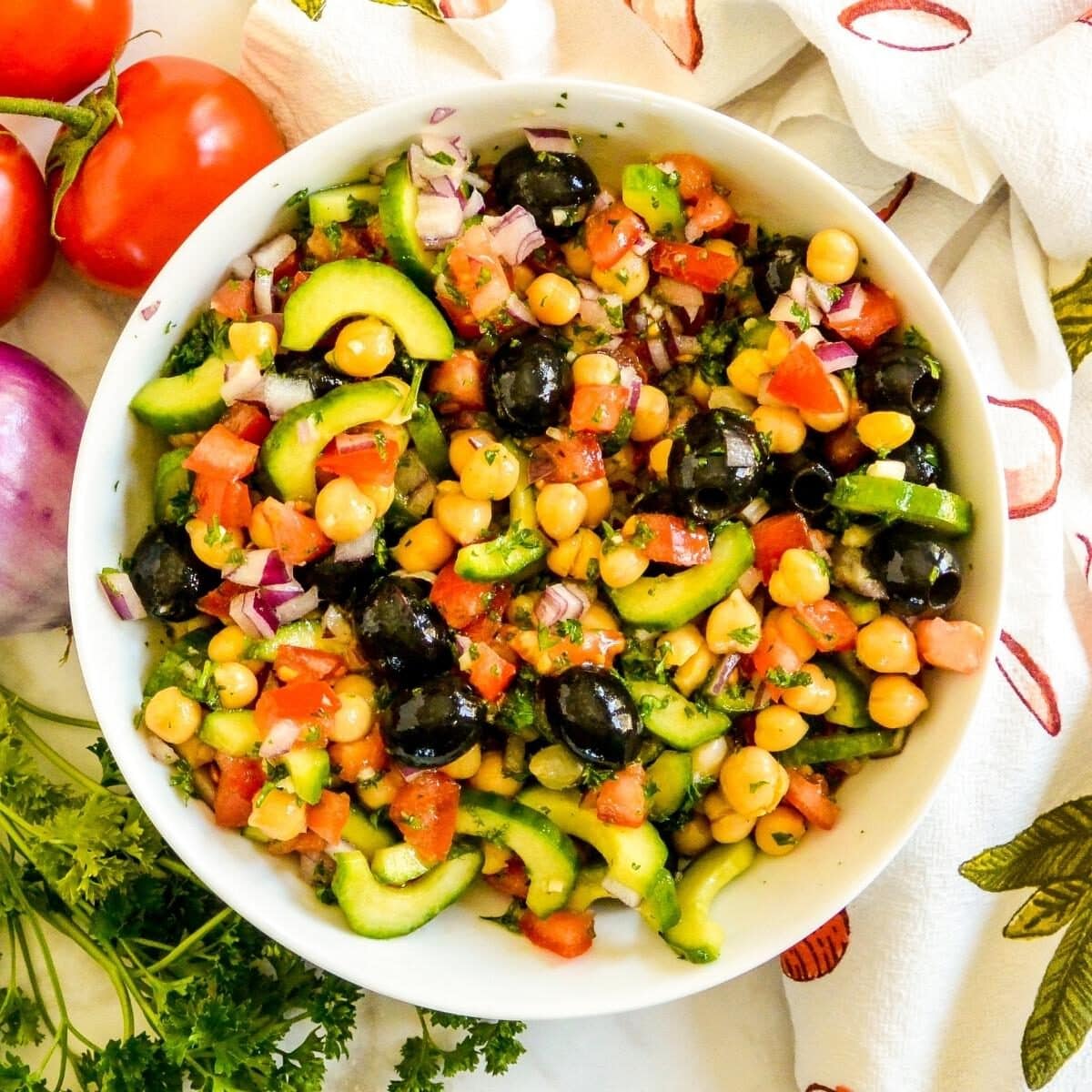 Bowl of Chickpea Cucumber Salad next to tomatoes, red onion, and fresh parsley.