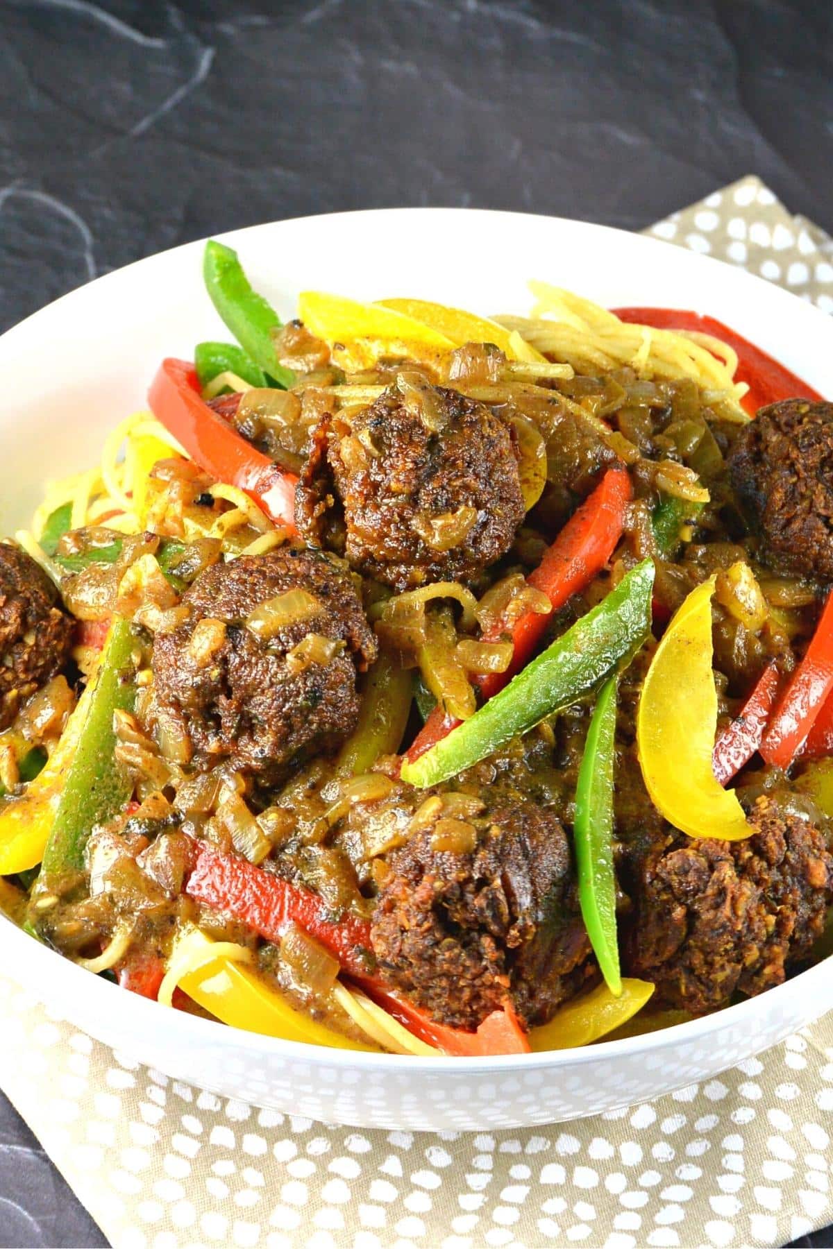 Side view of bowl of pasta topped with meatballs and red, yellow, and green bell pepper strips.