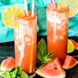 Glasses of Sparkling Watermelon Refresher garnished with slices of lime and surrounded by wedges of watermelon and sprigs of fresh mint.