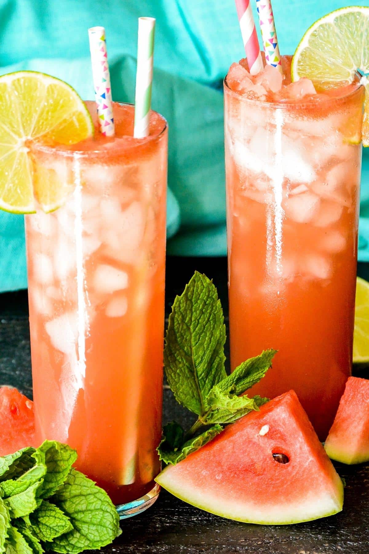 Two glasses of Sparkling Watermelon Refresher garnished with lime slices with watermelon wedges and sprigs of mint in the foreground.