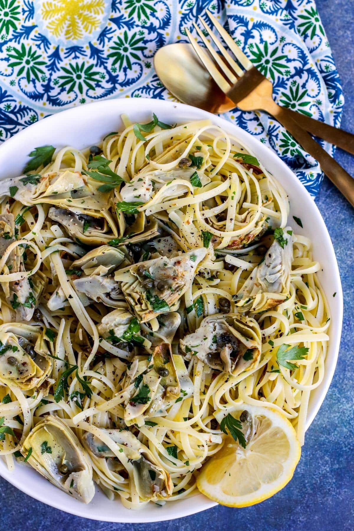 Plate of linguini with artichokes garnished with lemon slice with gold fork and spoon and printed napkin next to it.