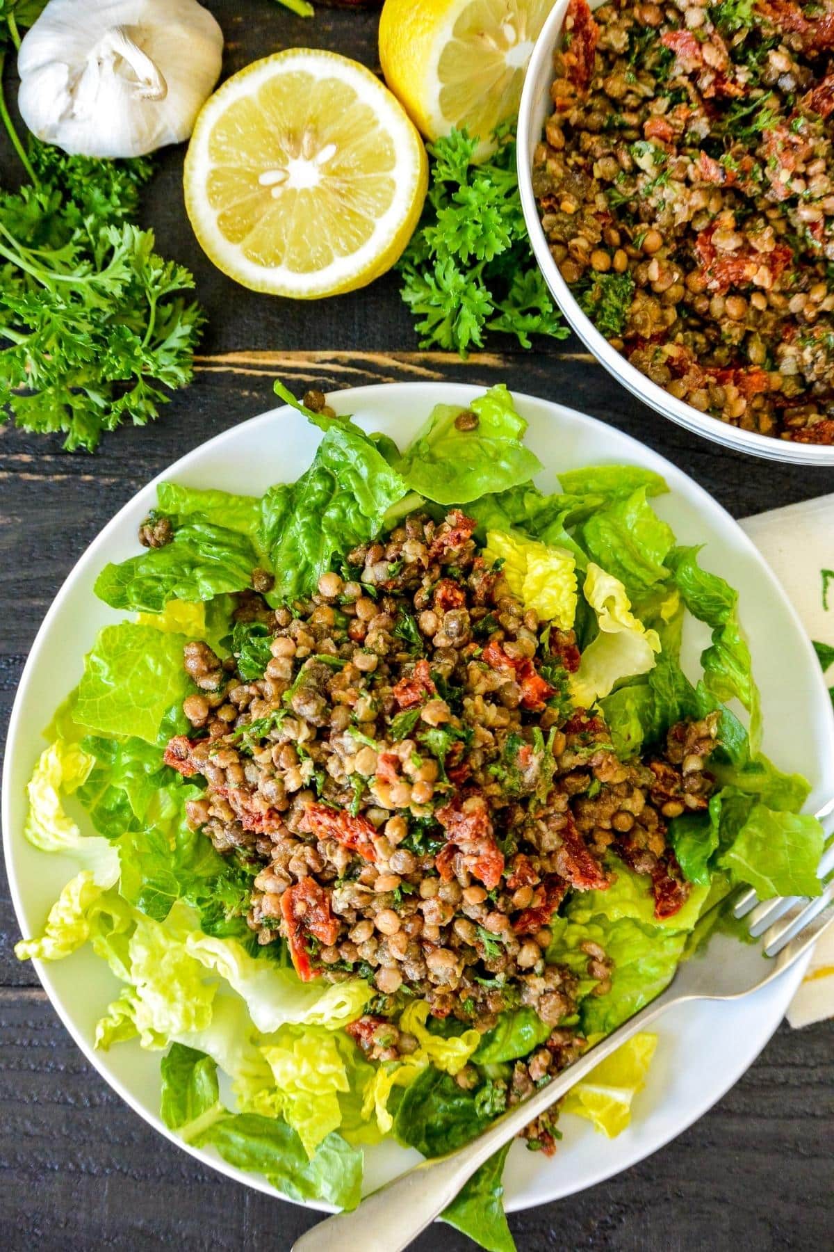 Plate with lentil salad on bed of chopped romaine lettuce with lemon, garlic, parsley, and salad bowl in the background.