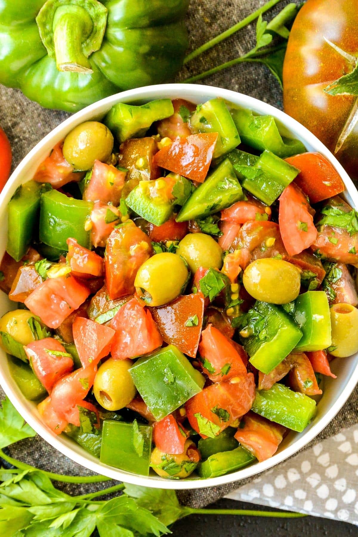 Overhead of a bowl of salad made with diced green bell peppers, heirloom tomatoes, green olives, and fresh parsley.