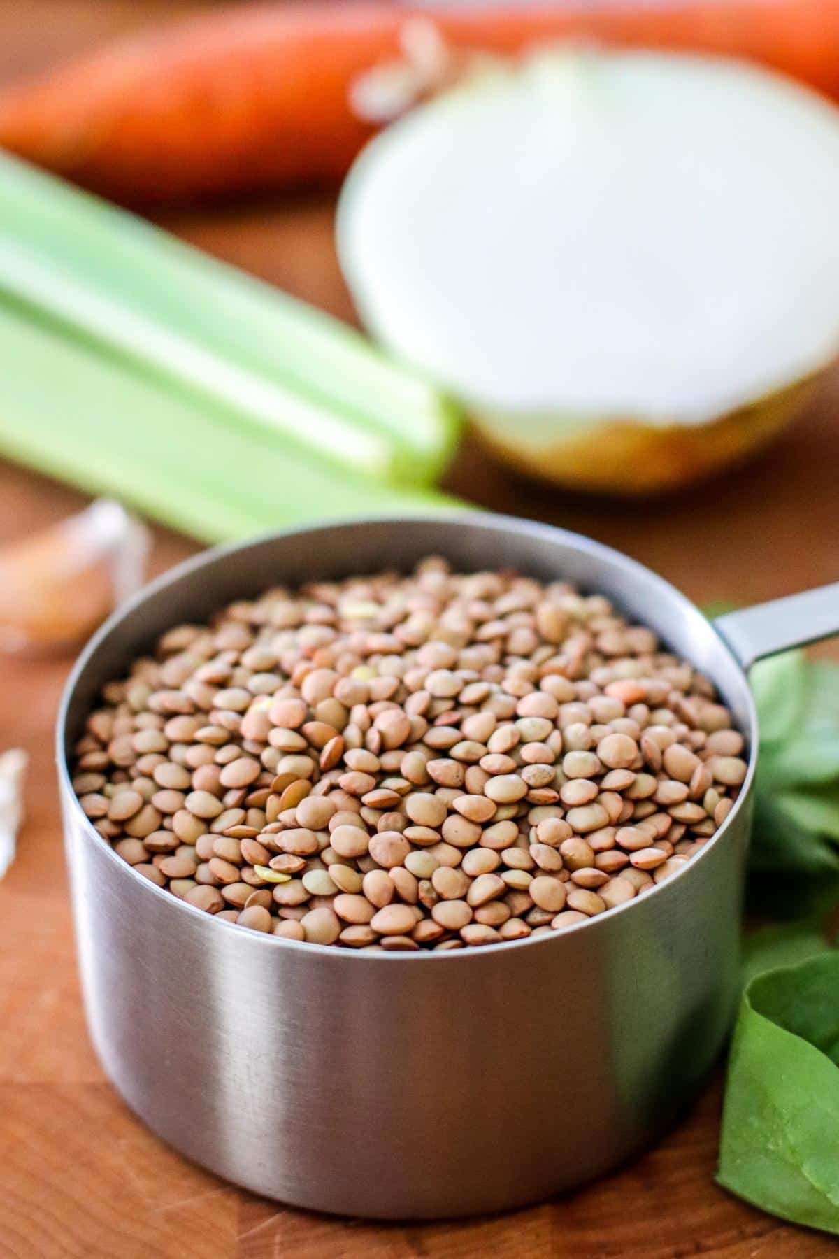 Cup of dry lentils on a cutting board with garlic cloves, celery ribs, carrot, half an onion, and some spinach.