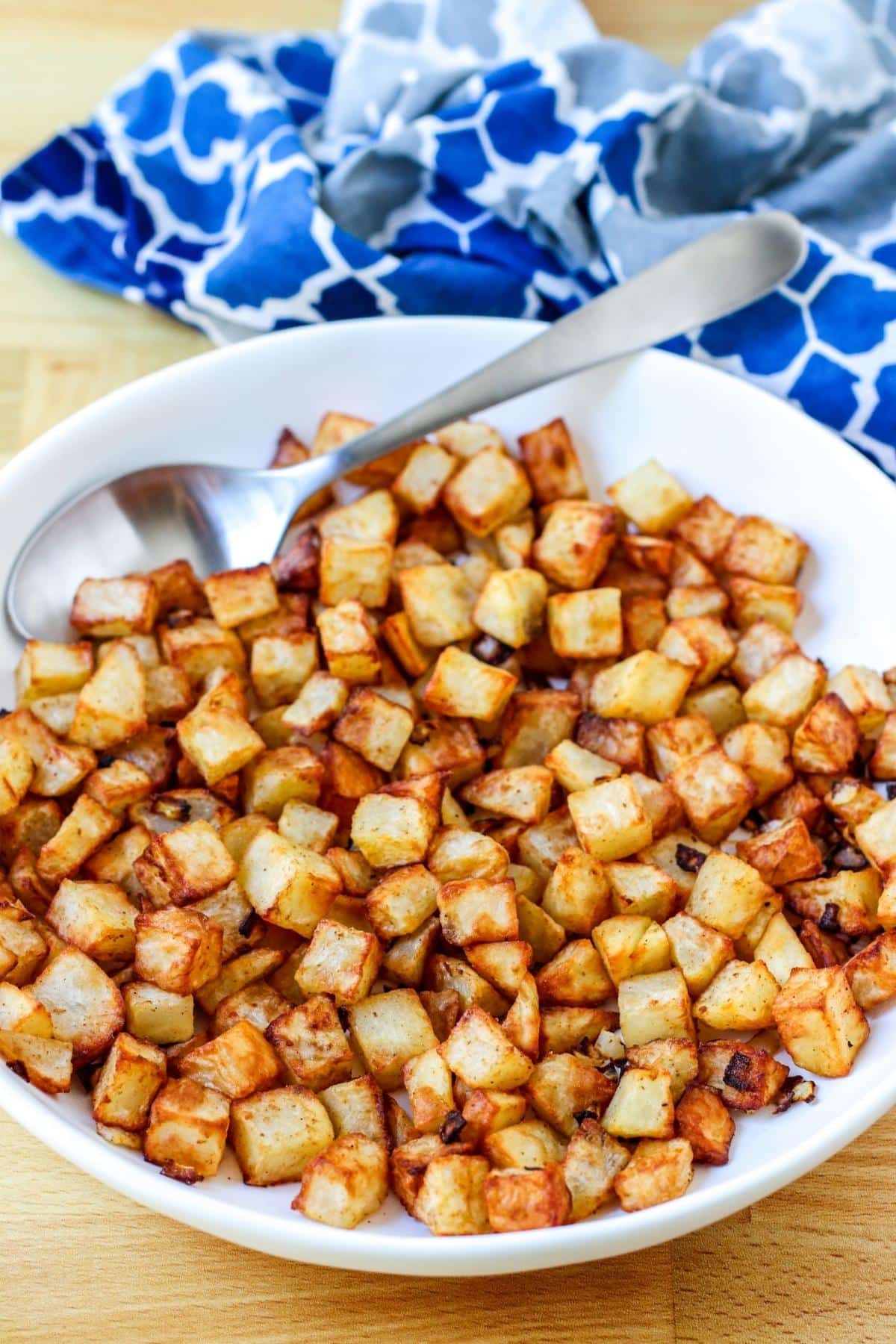 Home fries and a spoon in a shallow serving bowl with a blue patterned napkin in the background.
