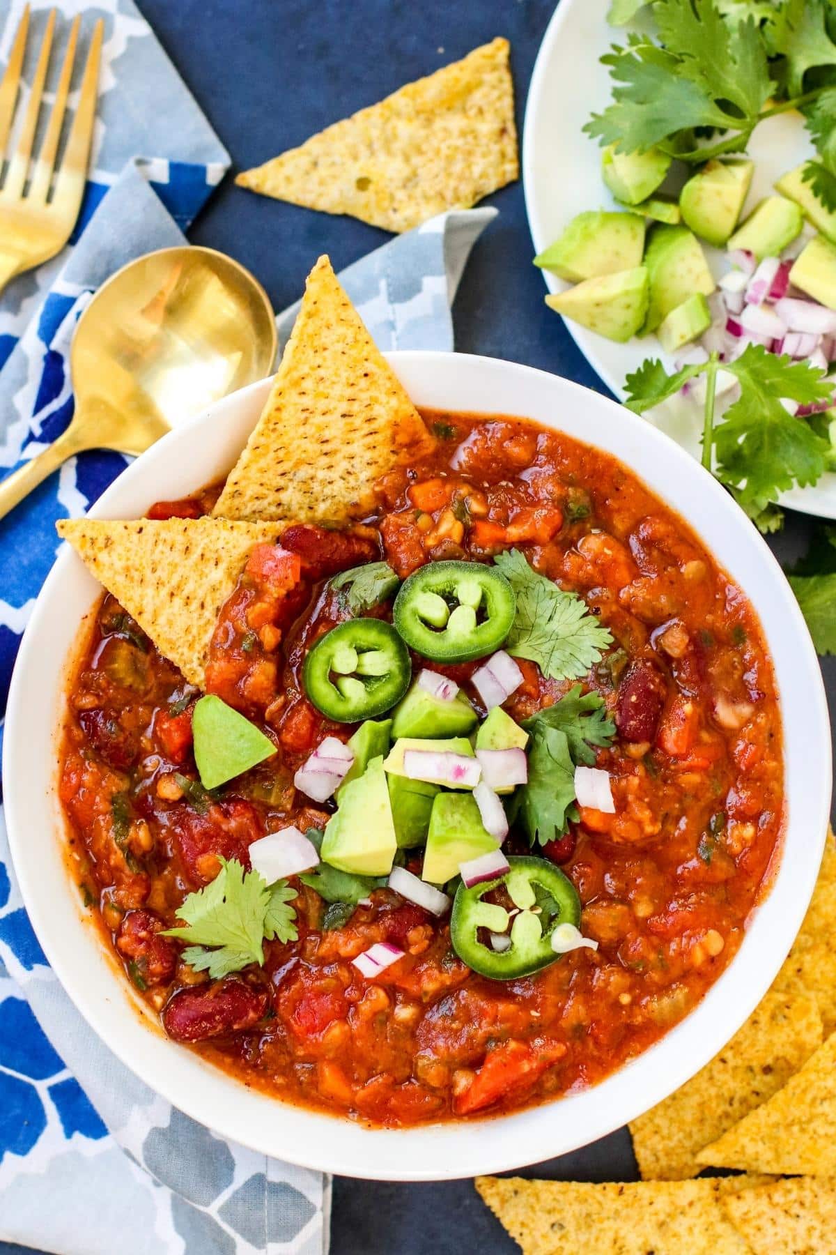 Bowl of red lentil chili garnished with jalapeno pepper slices, diced avocado, chopped red onion, and cilantro with tortilla chips and plate of toppings on the side.