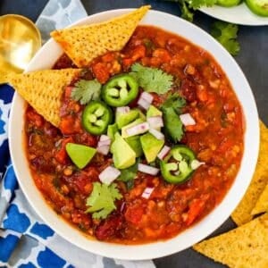 Bowl of chili topped with jalapeno pepper slices, red onions, and cilantro with tortilla chips on the side.