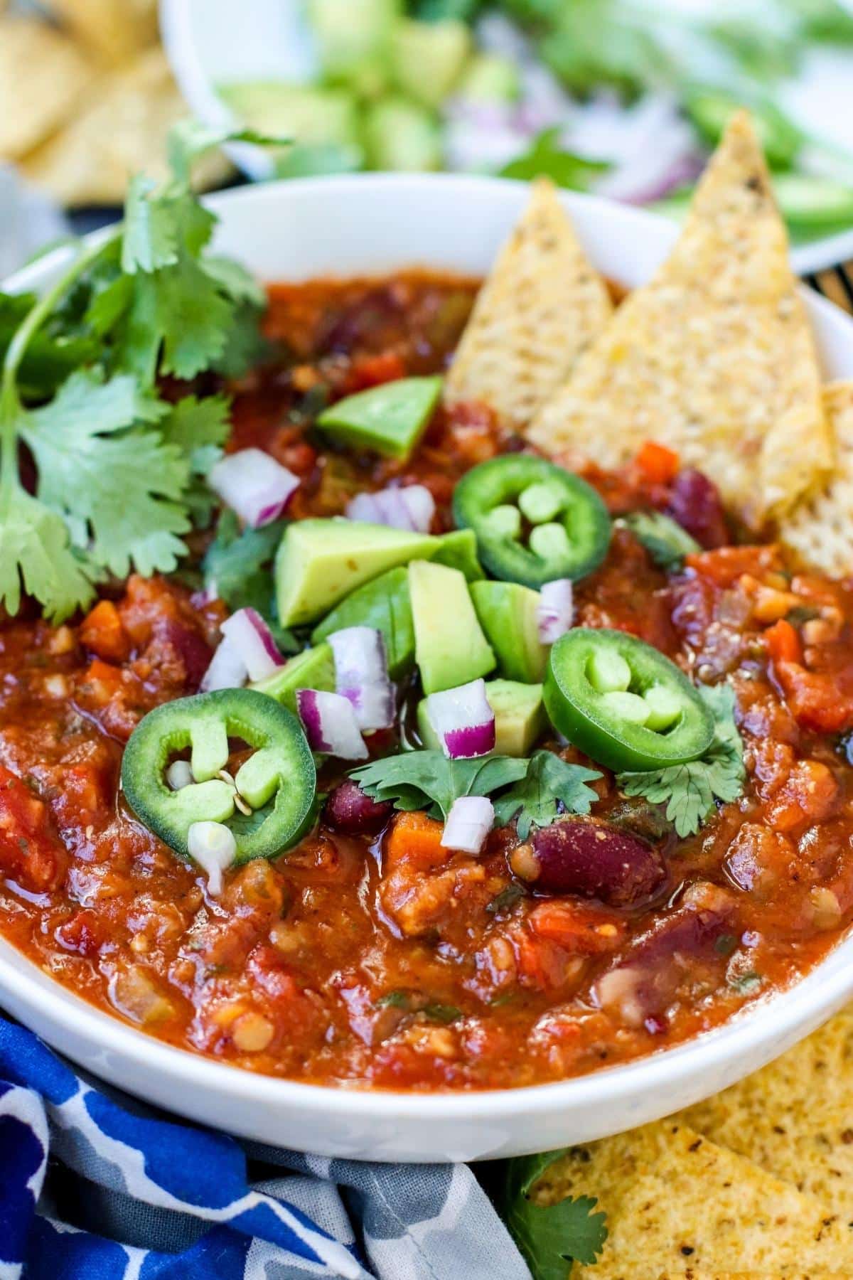 Close up of a bowl of chili topped with sliced jalapenos, avocado, red onions, and fresh cilantro with tortilla chips on the side of the bowl.