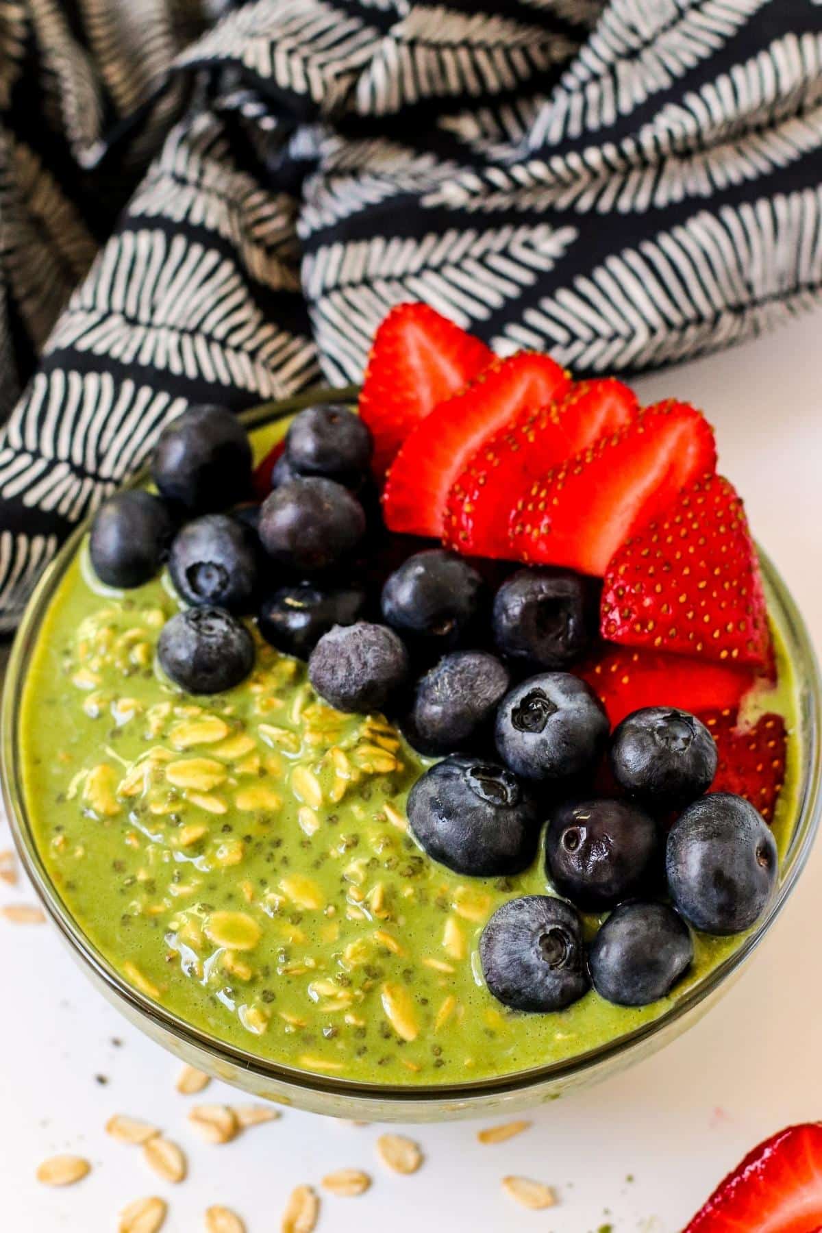 Bowl of green overnight oats topped with blueberries and sliced strawberries with blue and white napkin in the background.