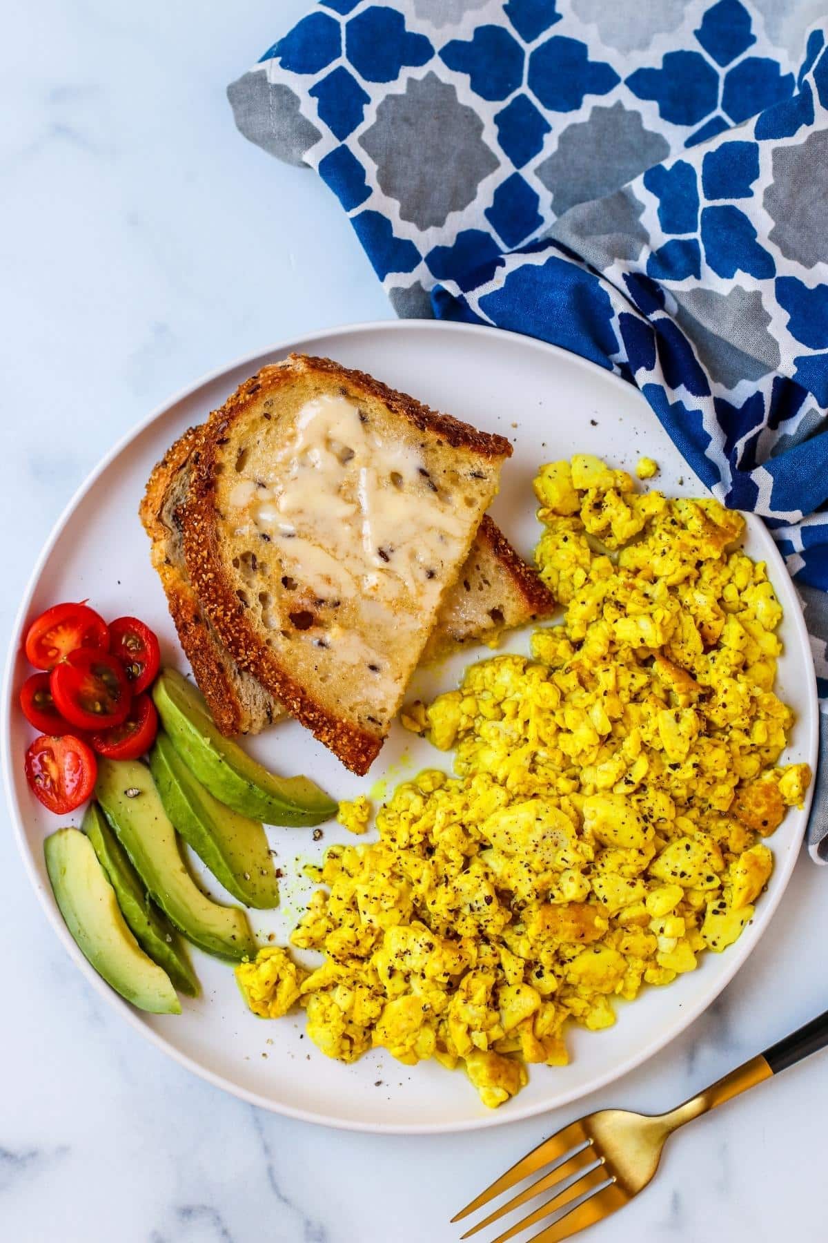Silken Tofu Scramble on a plate with toast and sliced avocado and tomatoes with gold fork and blue pattered napkin.