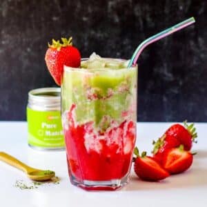 Strawberry Matcha Latte in a glass with a straw garnished with a fresh strawberry with jar of matcha powder and strawberries next to it.