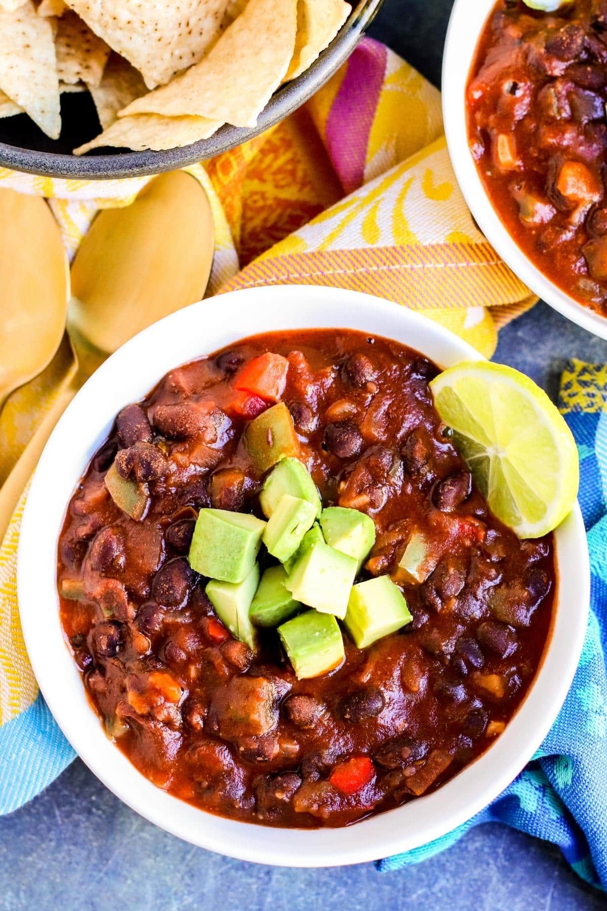 Bowl of vegan black bean chili topped with avocado and a slice of lime with a bowl of tortilla chips on the side.