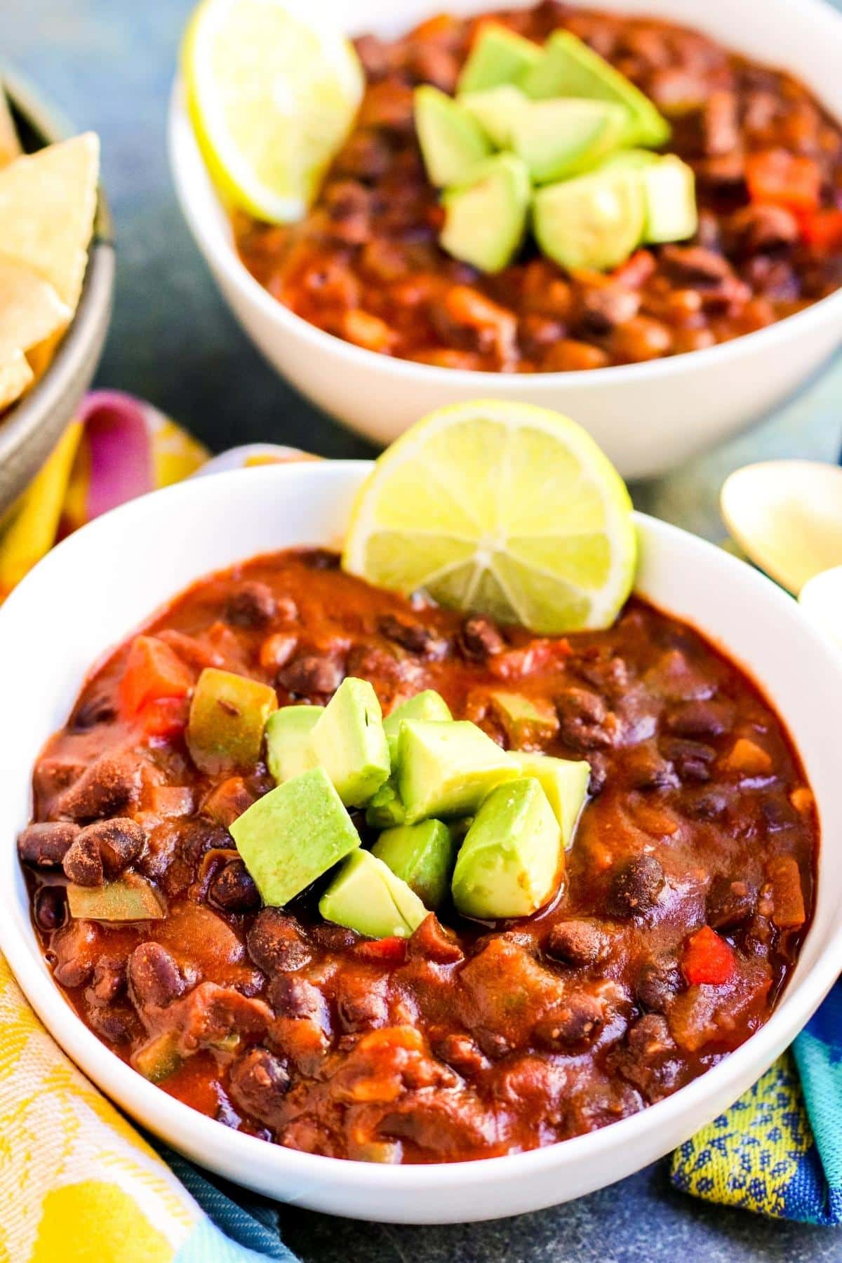 Two bowls of chili topped with avocado and garnishes with slices of lime.