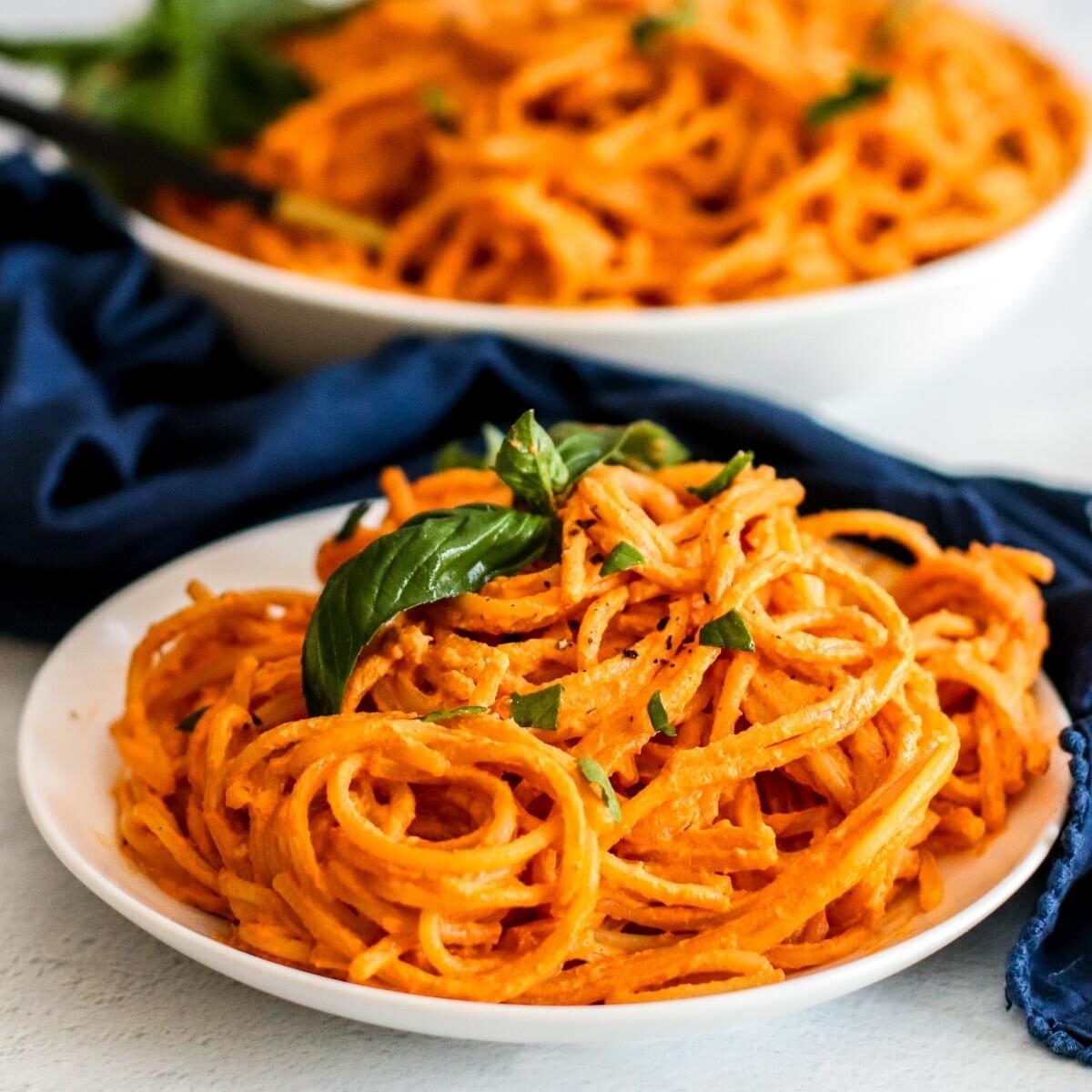 Plate of spaghetti with creamy red sauce garnished with sprig of fresh basil with serving bowl in the background.