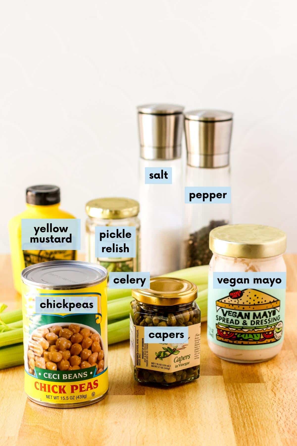 Ingredients for this recipe with text labels on a cutting board: Can of chickpeas, bottle of yellow mustard, jar of pickle relish, jar of capers, bunch of celery, jar of vegan mayo, and salt and pepper grinders.