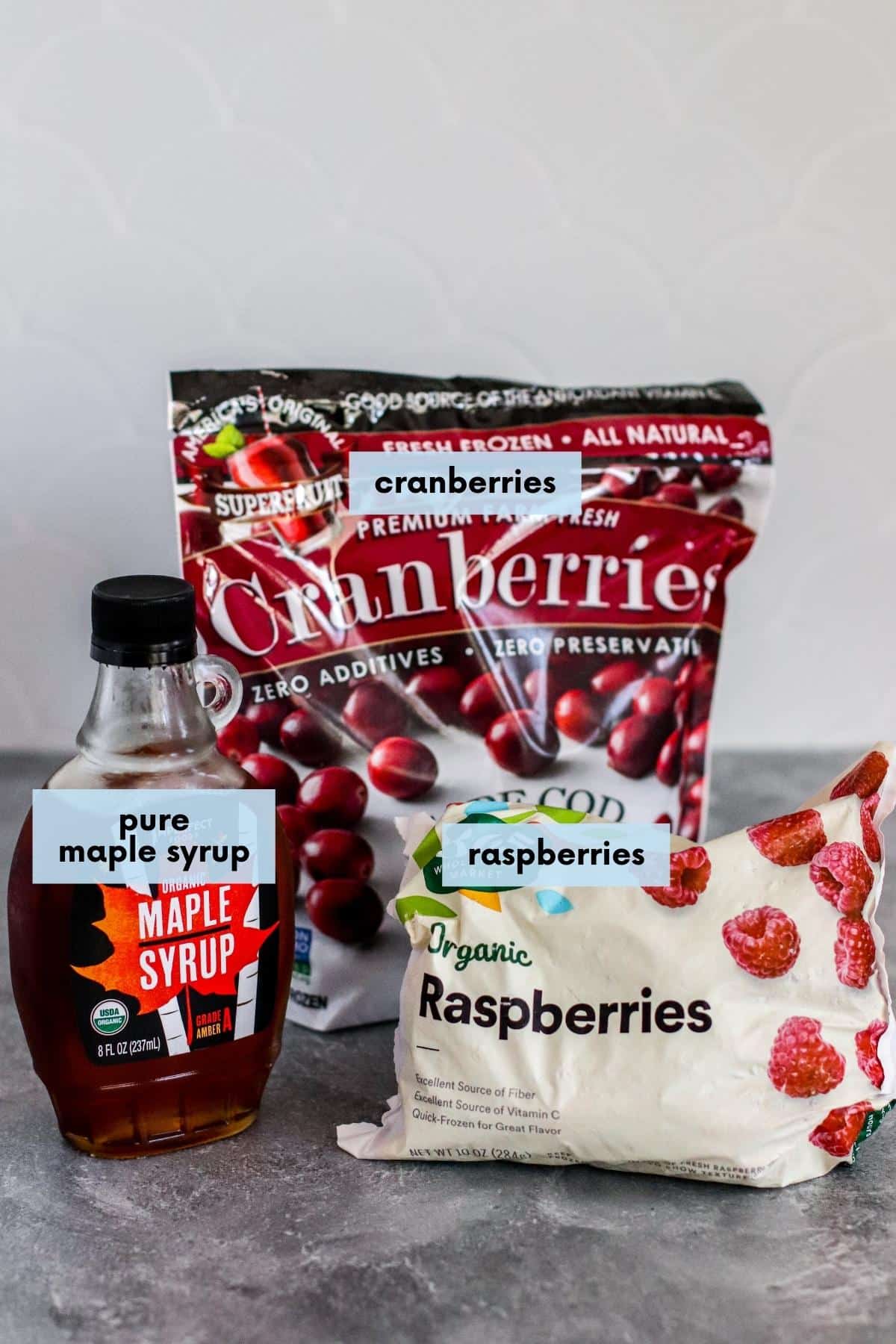 Ingredients on a countertop: Bottle of pure maple syrup, bag of frozen cranberries, and bag of frozen raspberries.