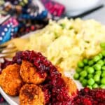 Dinner plate with tofu meatballs topped with cranberry raspberry sauce, mashed potatoes, and peas with bowl of sauce in the background.