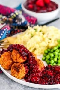 Dinner plate with tofu meatballs topped with cranberry raspberry sauce, mashed potatoes, and peas with bowl of sauce in the background.