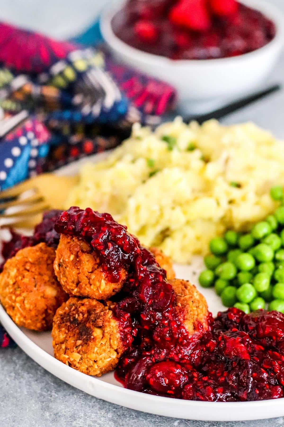 Dinner plate with tofu meatballs topped with cranberry raspberry sauce, mashed potatoes, and peas with bowl of sauce in the background.