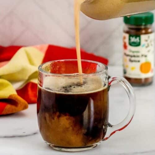 Pumpkin creamer being poured into a glass mug of black coffee with a bottle of pumpkin pie spice in the background.