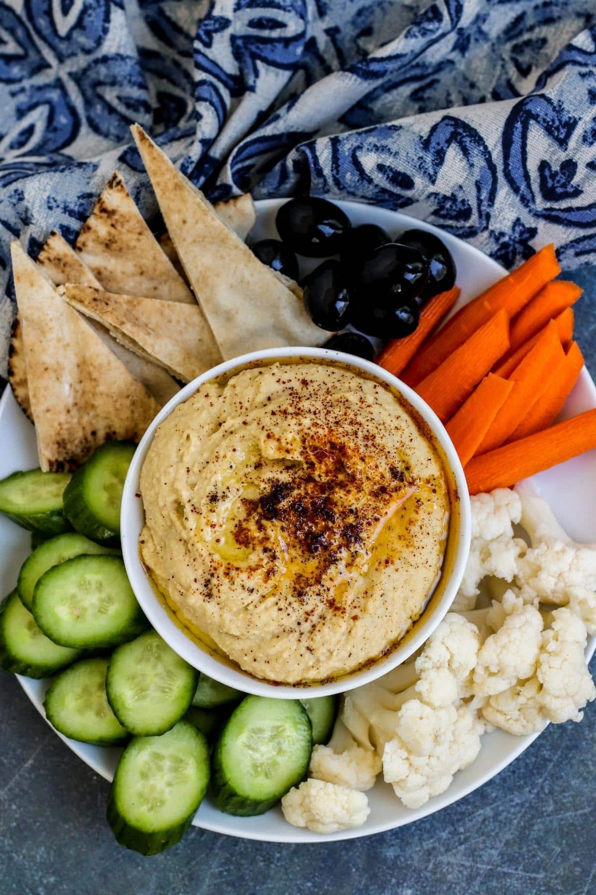 Platter of pita bread triangles, black olives, carrot sticks, cauliflower florets, and sliced cucumbers with a bowl of hummus in the middle.