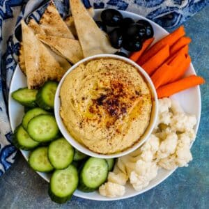Bowl of hummus on a platter with raw vegetables and pita bread triangles.