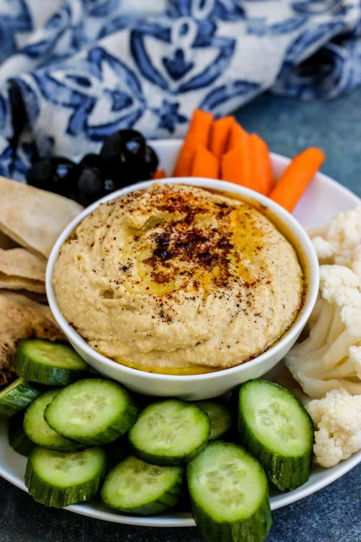 A bowl of hummus served with cucumbers, cauliflower, carrots, olives, and pita bread.