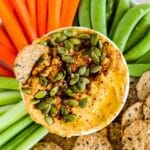 Bowl of pumpkin hummus garnished with pumpkin seeds and spices on a platter of celery, carrots, sugar snap peas, and crackers.