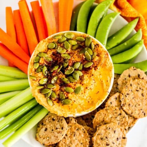 Bowl of pumpkin hummus garnished with spices and pumpkin seeds and surrounded by raw vegetables and crackers.