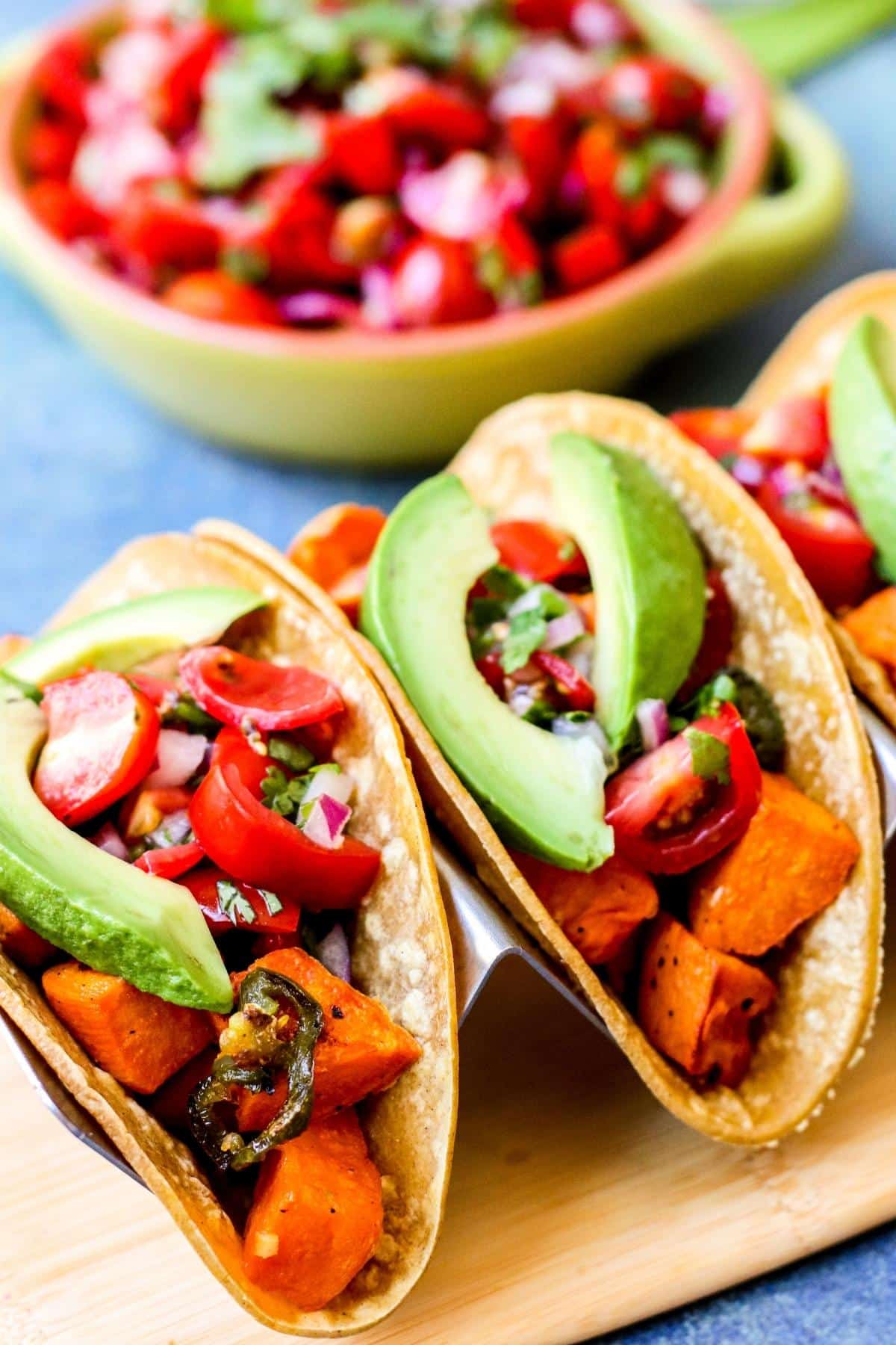 Tacos filled with roasted cubes of sweet potatoes and slices of jalapeño topped with fresh salsa and slices of avocado.