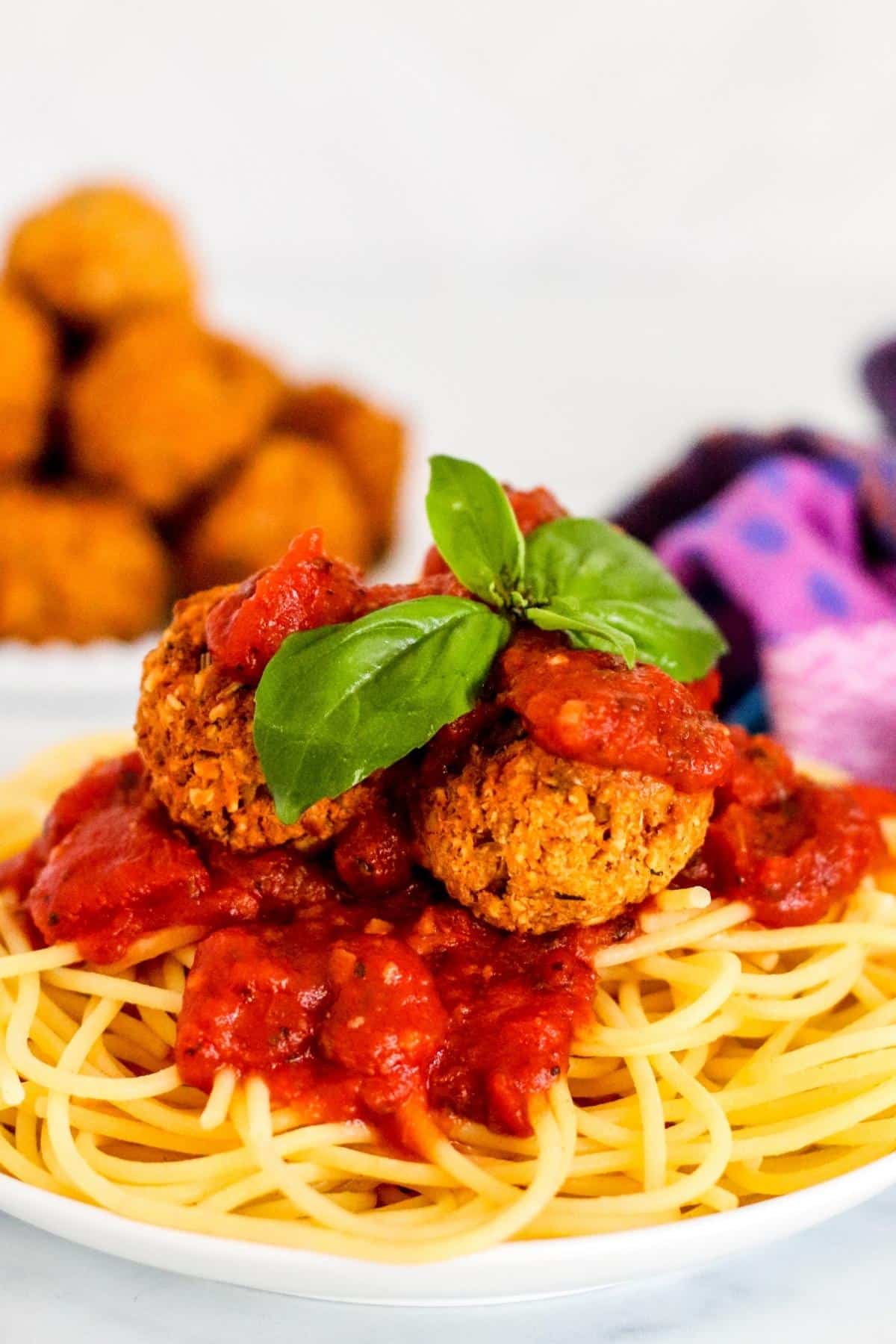 Plate of spaghetti topped with red sauce, tofu meatballs, and fresh basil with platter of meatballs in the background.