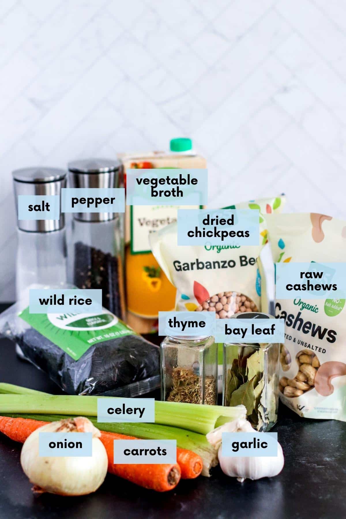 Labeled ingredients on a kitchen counter: Onion, carrots, celery, garlic, wild rice, salt and pepper, vegetable broth, dried thyme, bay leaf, dried chickpeas, and raw cashews.