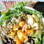Bowl of arugula, quinoa, black beans, roasted butternut squash, chopped apples, and pumpkin seeds with a drizzle of dressing and text overlay Butternut Squash Buddha Bowls.