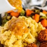 Pouring onion gravy over mashed potatoes on a dinner plate with roasted vegetables and tofu meatballs.