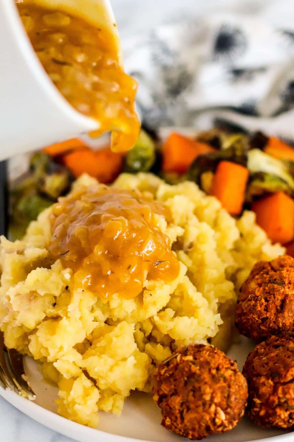 Pouring onion gravy over mashed potatoes on a dinner plate with roasted vegetables and tofu meatballs.