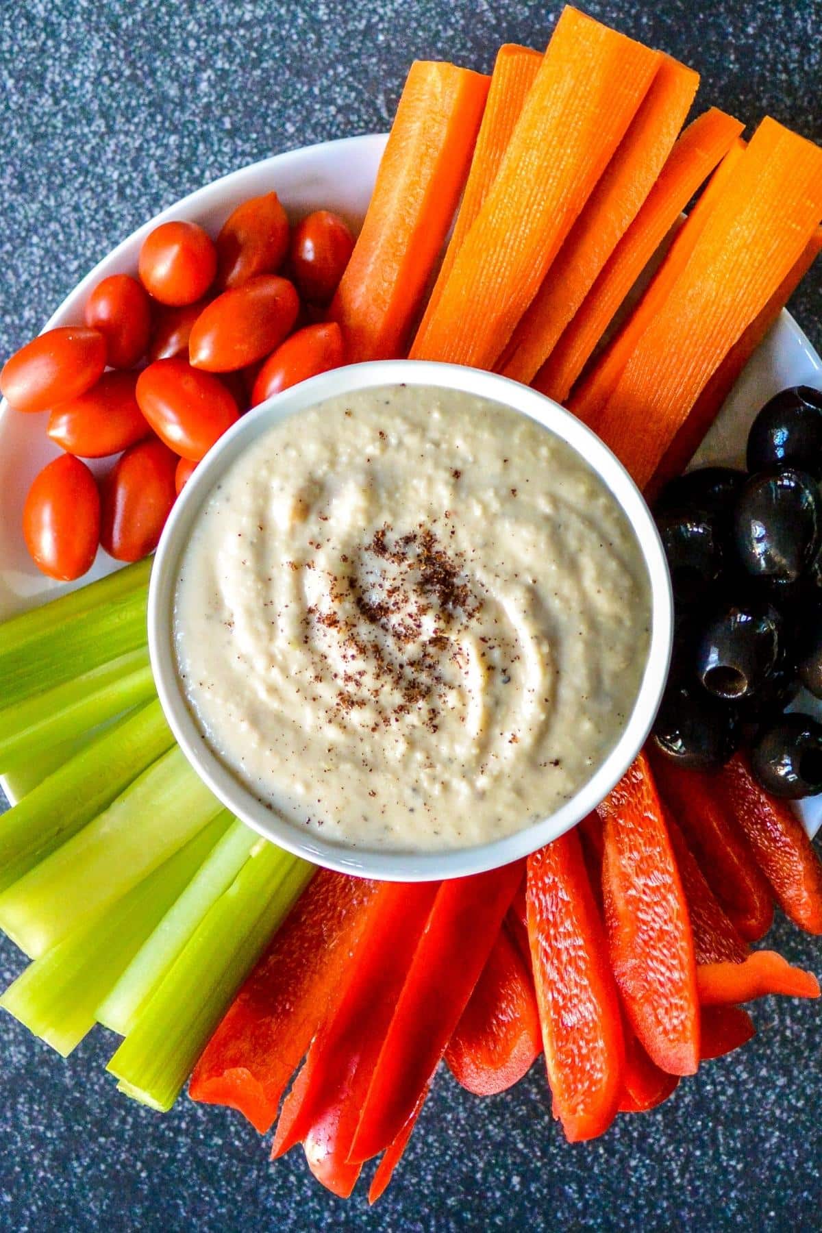 Bowl of dip on a platter with grape tomatoes, carrot sticks, black olives, red pepper strips, and celery sticks.