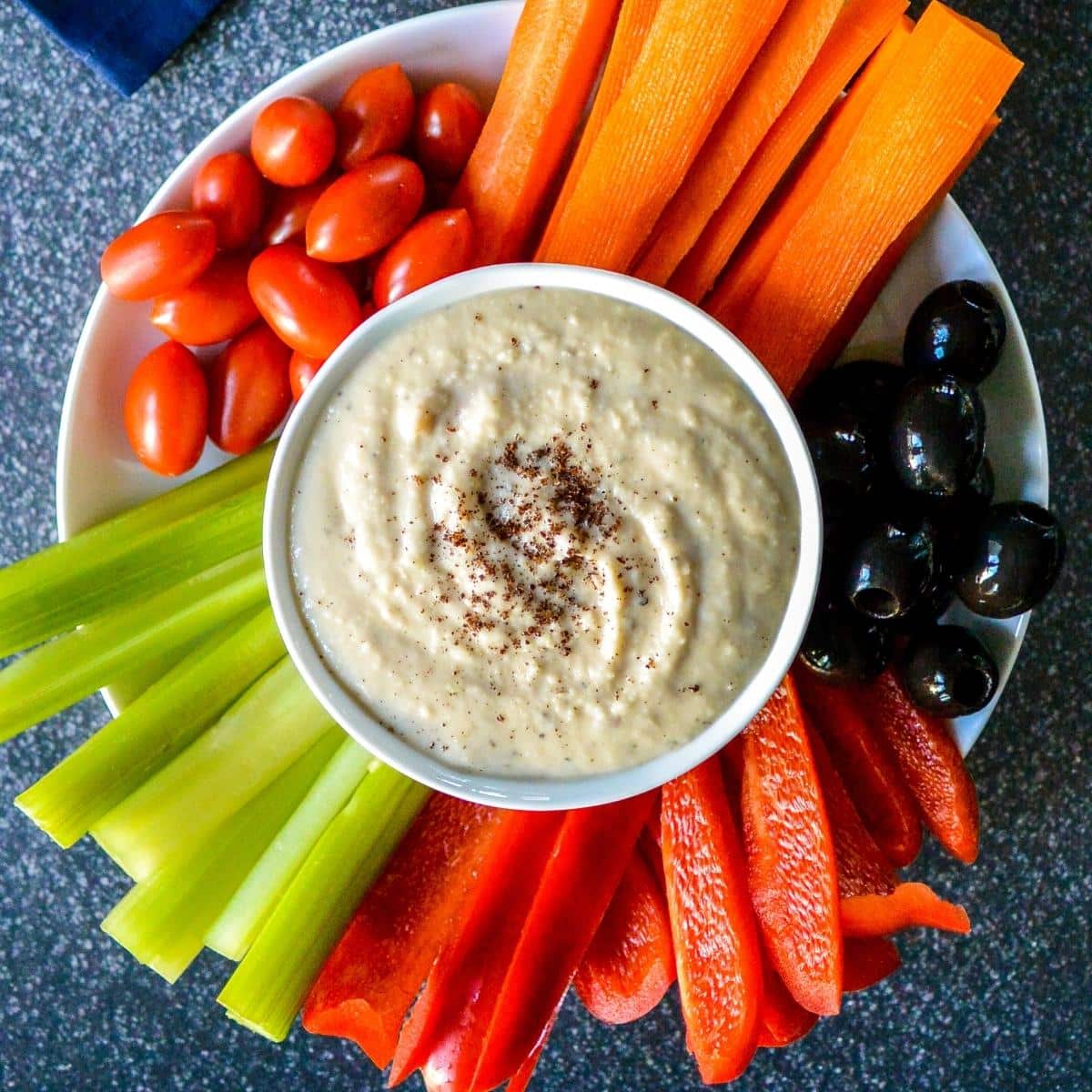 Bowl of dip on a platter with grape tomatoes, carrot sticks, black olives, red pepper strips, and celery sticks.