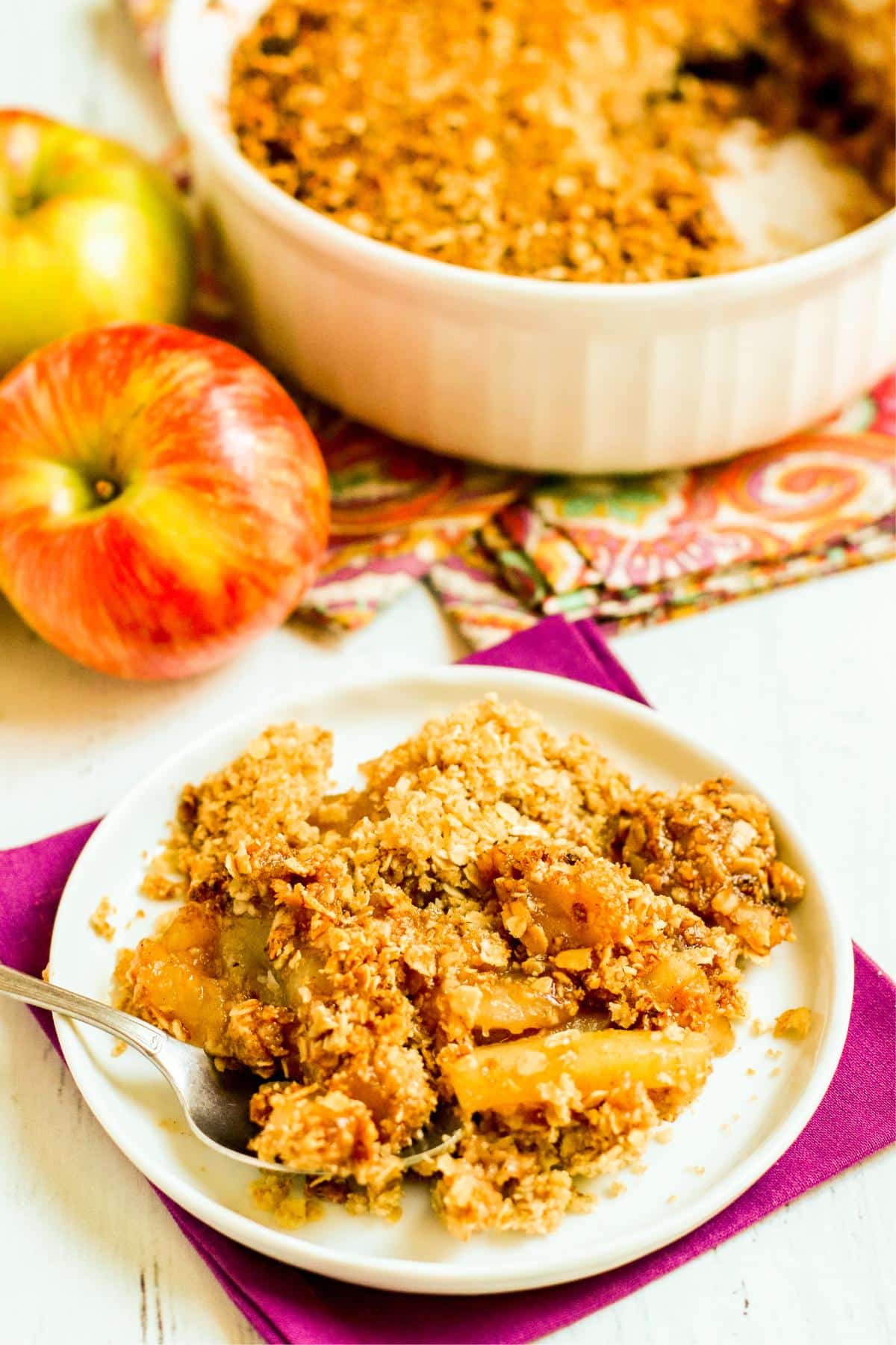 Plate with a spoon in a serving of vegan apple crisp with apples and baking dish in the background.
