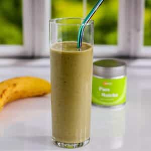 Banana Matcha Smoothie in a glass with a metal straw with a banana and container of matcha in the background.