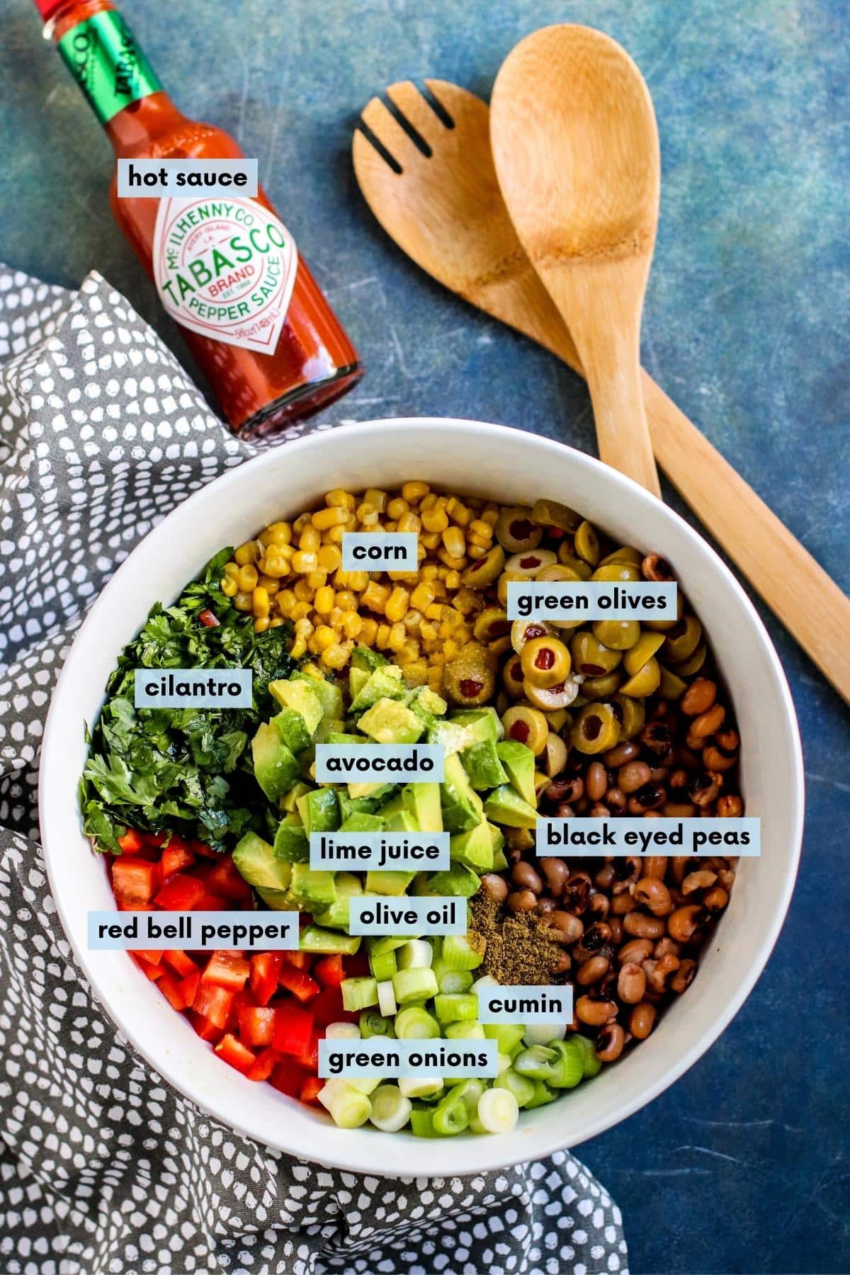Bottle of hot sauce next to a large bowl with the dip ingredients in it and salad utensils on the side.