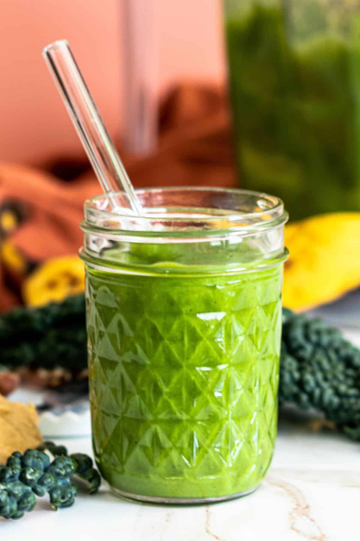 Glass of Peanut Butter Green Smoothie with a glass straw in it with kale leaves, a banana, and the smoothie pitcher in the background.