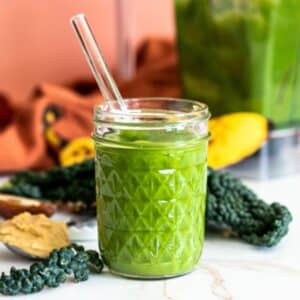 Smoothie with a glass straw with ingredients and blender pitcher in the background.