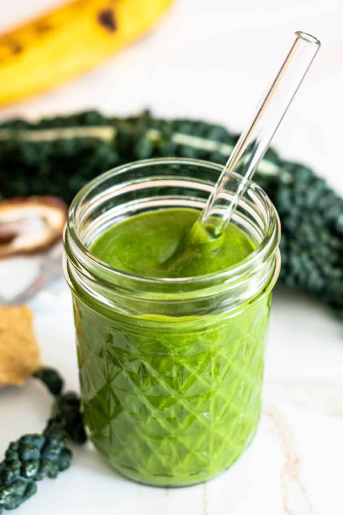 Green smoothie with a glass straw with kale leaves and a ripe banana in the background.